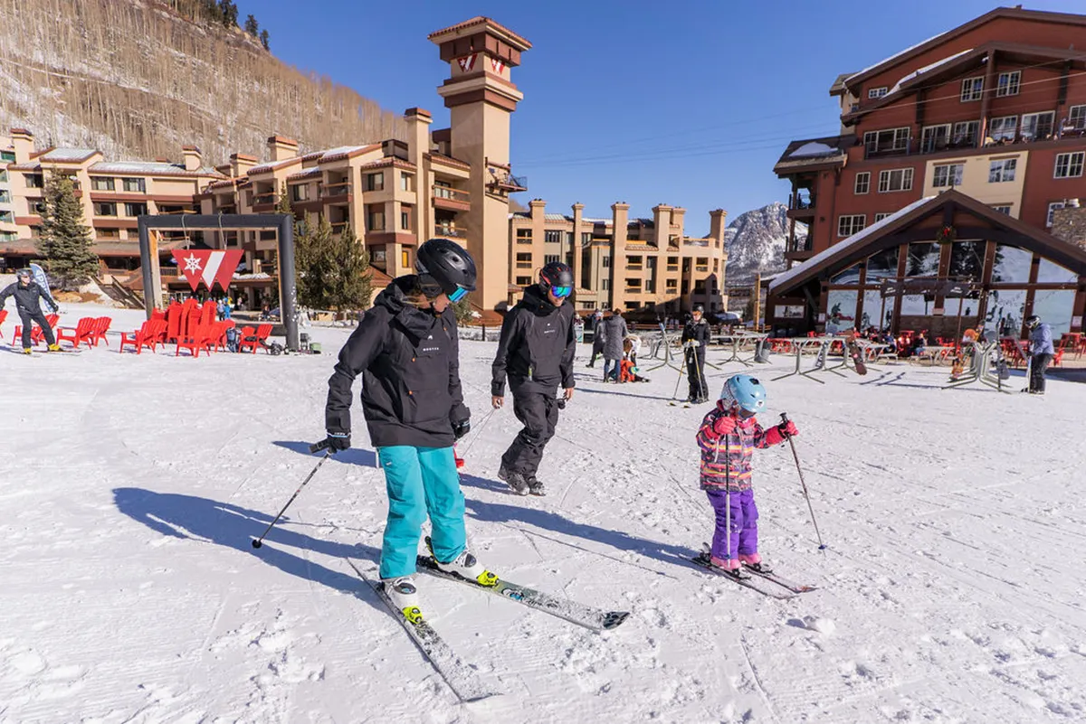 A family is captured skiing together on snow-covered terrain with the resort buildings in the background. The scene is bright and playful, emphasizing shared excitement and the joy of learning and playing together in the mountains.