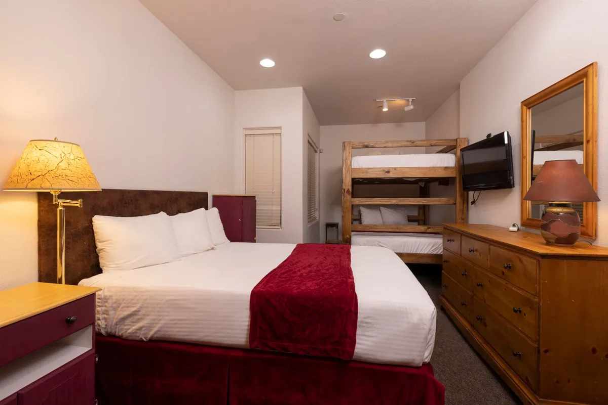 A family-friendly bedroom with a queen bed featuring a red throw and an adjacent bunk bed, accompanied by a wooden dresser, a uniquely designed lamp, and a television. The space feels cozy, ideal for both families and couples seeking privacy and comfort.