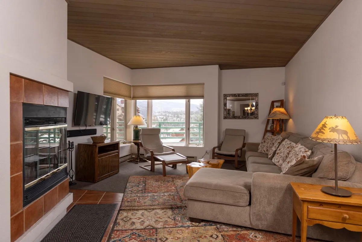The image showcases a cozy living room with a crackling fireplace as its centerpiece, a plush sofa arranged for comfort, a coffee table ideal for gatherings, and a decorative lamp that casts a warm glow. Through a large window, natural light pours in, hinting at breathtaking mountain vistas beyond.