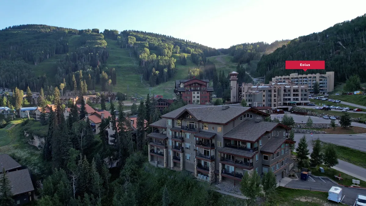 A view of residential resort buildings with a clear mountain backdrop and a parking area in the foreground. The scene includes covered parking spaces and highlights the ease of access and shelter from the elements, adding to the comfort of resort living.