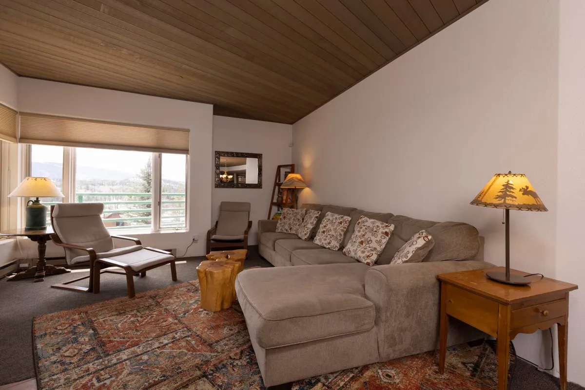 A spacious living area with a large sectional sofa faces floor-to-ceiling windows, which flood the room with natural light and frame dramatic mountain vistas. A wooden side table and soft lamps complement the comfortable, inviting setting—ideal for unwinding, sharing stories, or enjoying the changing scenery throughout the day.