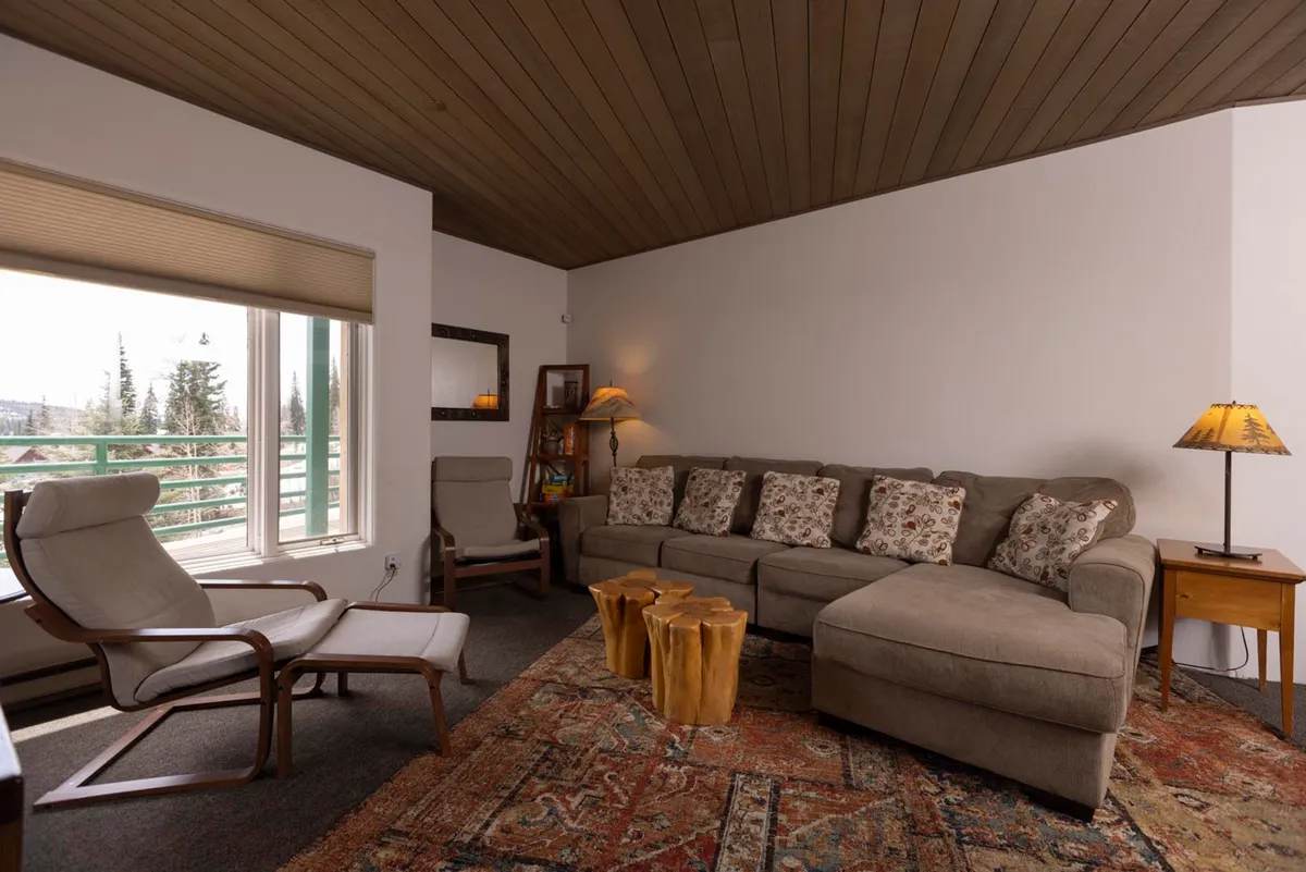 Light pours into the living room through expansive windows, illuminating a plush sectional sofa and a wooden coffee table set for board games or conversation. The view outside shows treetops and hints of mountains, inviting you to watch the sunrise or settle in for a cozy gathering. The space exudes comfort, making it perfect for morning coffee or an evening with friends.