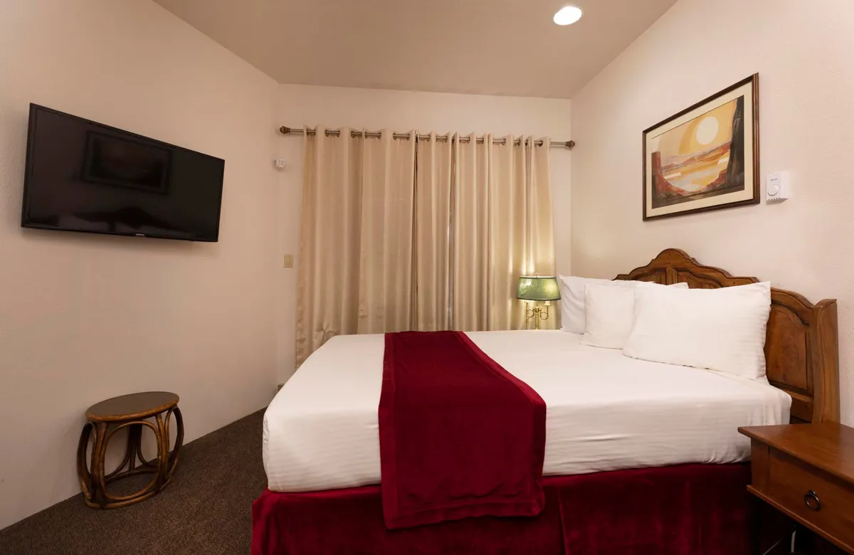 A spacious bedroom featuring a queen bed adorned with a red throw, a wooden bedside table, large flat-screen TV, and soft curtains. Warm lighting and plush bedding create an inviting, restful atmosphere, perfect for unwinding after a busy day.