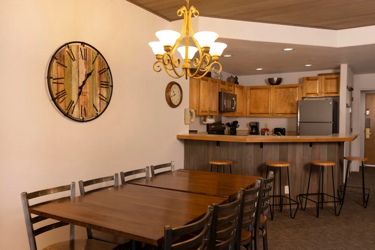 A warm, welcoming dining nook featuring a dining table under a chandelier, a visible clock, and a kitchen bar. The area is set for family meals and social gatherings, with ample natural light and a clear connection to the kitchen.