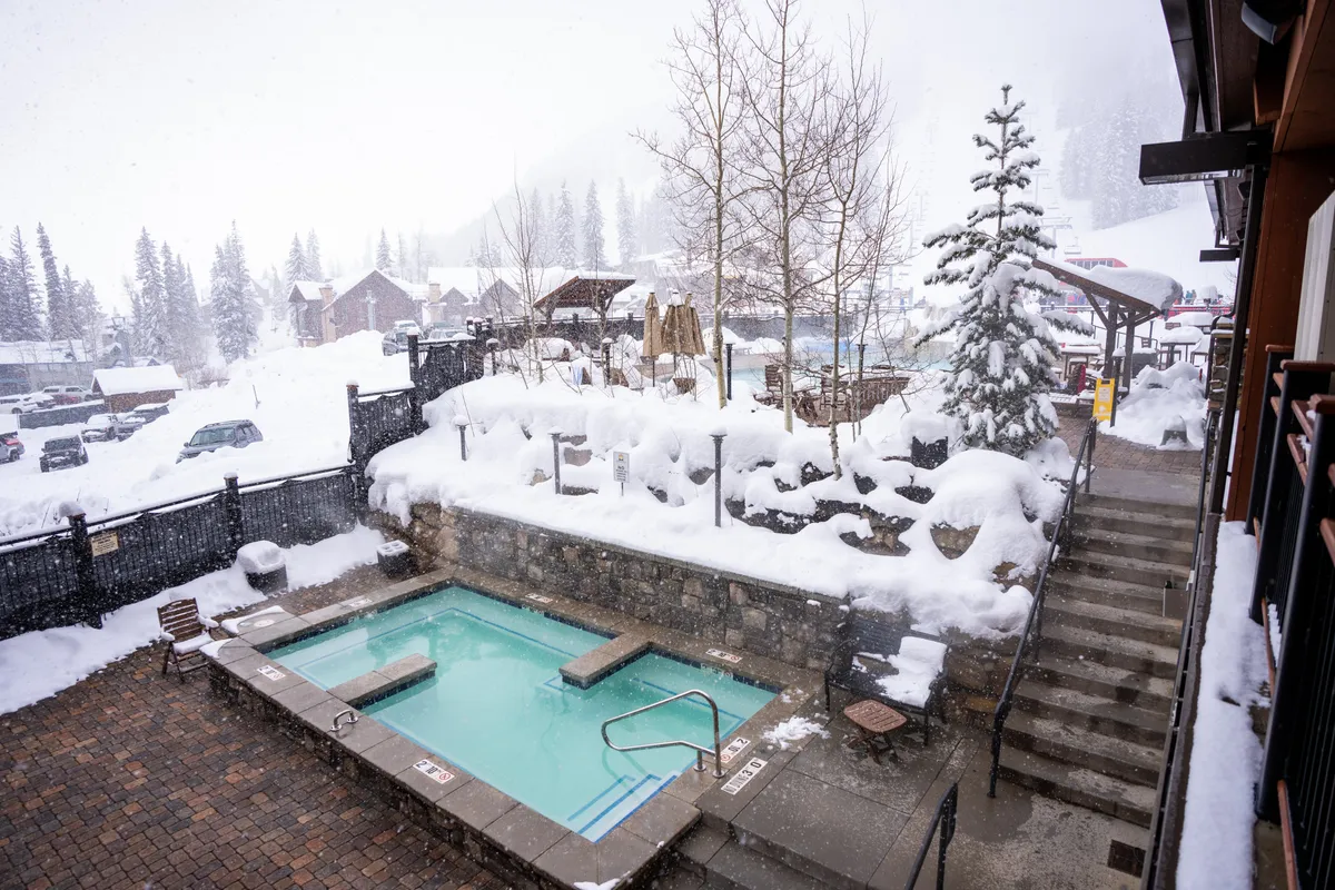 A steaming hot tub set outdoors, surrounded by a snow-covered landscape and tall pine trees. A sturdy stone wall adds rustic charm, while steam rises from the bubbling water. The scene evokes a sense of cozy relaxation and winter comfort, ideal after active days on the slopes.