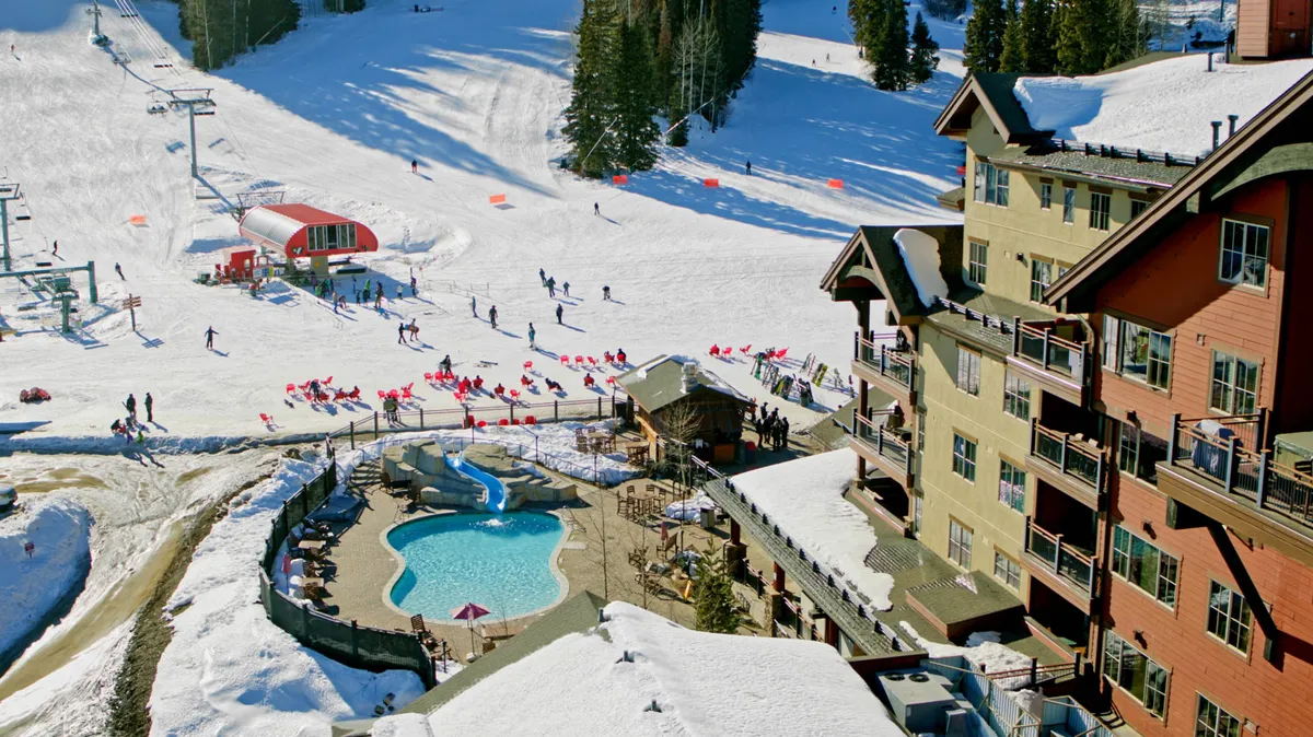 A lively winter scene outside the lodge, showcasing ski slopes dotted with skiers and snowboarders. The pool area is visible, steam rising against the snowy backdrop, and the lodge’s distinctive architecture frames the action. In the distance, mountains provide a dramatic setting for outdoor fun and relaxation alike.